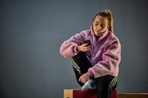 Fi in a furry pink jacket sits on a box looking at a phone